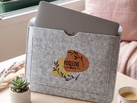 Housse d'ordinateur en feutrine grise avec motif coloré Positive Vibes posée sur un bureau en bois clair avec un PC et une tasse.