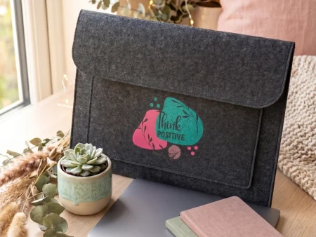 Housse d'ordinateur en feutrine grise avec motif coloré Think Positive posée sur un bureau blanc avec une tasse de café et un stylo doré.