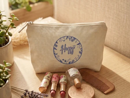 Pochette en coton écru avec logo Happy bleu posée debout sur une coiffeuse avec des rouges à lèvres et des fleurs séchées.